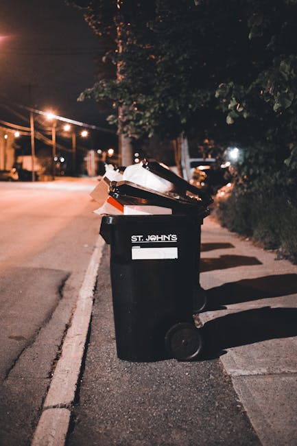 A row of large wheelie bins positioned along a sidewalk against a brick wall, with green and blue bins prominently visible in the foreground and red bins further back. The bins are made of plastic wit
